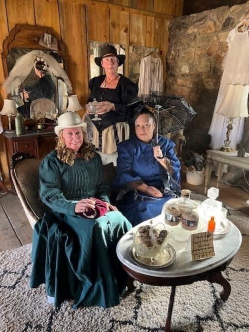 Three women in vintage dresses and hats sit in a rustic room, with a table set for tea and an umbrella nearby.