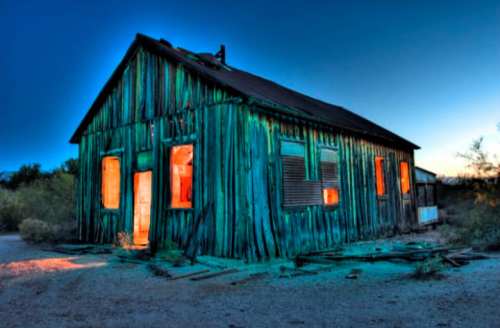 A weathered wooden house at dusk, illuminated windows glowing against a deep blue sky.