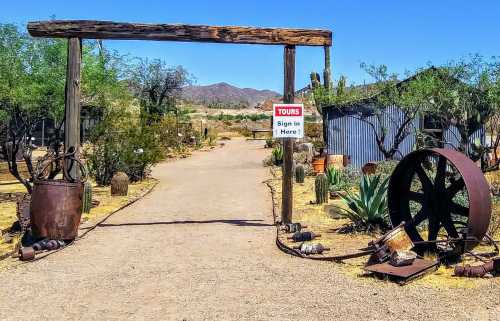 Entrance to a desert garden with a wooden arch, sign for tours, and rustic decor including a large wheel and pots.