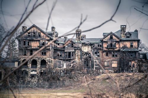 A dilapidated, abandoned mansion with broken windows and overgrown vegetation, set against a cloudy sky.