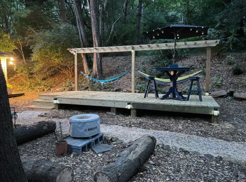 A wooden deck in a forested area features a table, chairs, a hammock, and an umbrella, surrounded by trees and logs.