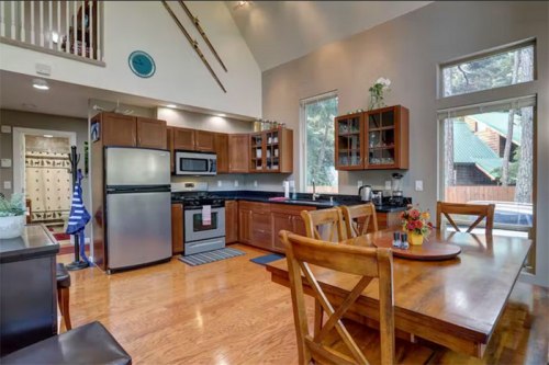 Bright kitchen with wooden cabinets, stainless steel appliances, and a dining table, featuring large windows and natural light.