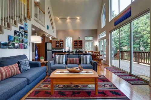 Bright living room with large windows, cozy sofas, a wooden coffee table, and a view of a deck and trees outside.