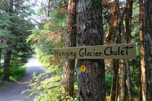 A wooden sign pointing to "Hanging Glacier Chalet" amidst trees along a gravel path.