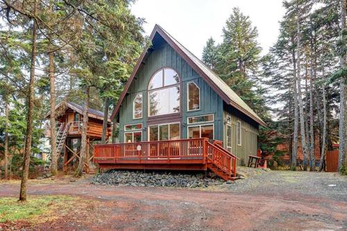 A modern A-frame cabin surrounded by trees, featuring a large deck and large windows.