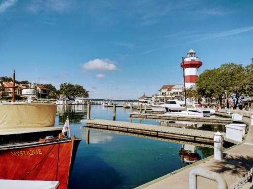 A serene marina scene with boats, a red and white lighthouse, and clear blue skies reflecting on calm waters.