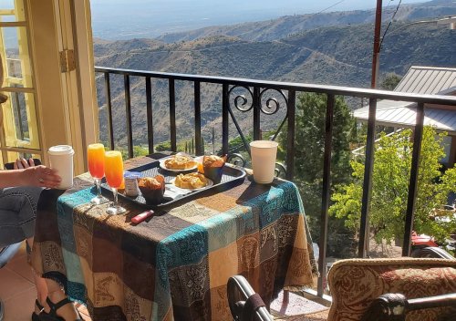 A scenic breakfast table with pastries and drinks, overlooking a mountainous landscape. Sunlight brightens the scene.