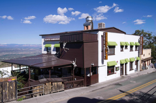 A two-story restaurant with green awnings, featuring a patio, set against a scenic landscape and blue sky.