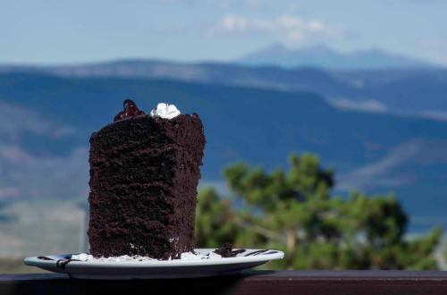 A slice of rich chocolate cake with whipped cream, set against a scenic mountain backdrop.