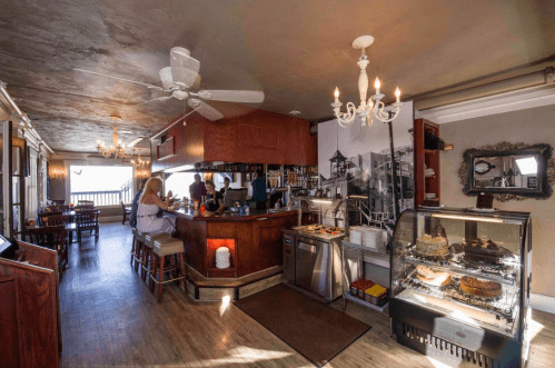 Cozy café interior with a bar, chandelier, and display case of desserts, featuring a view of the outdoors.