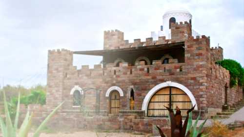 A stone castle with turrets and arched windows, surrounded by greenery and a cloudy sky.