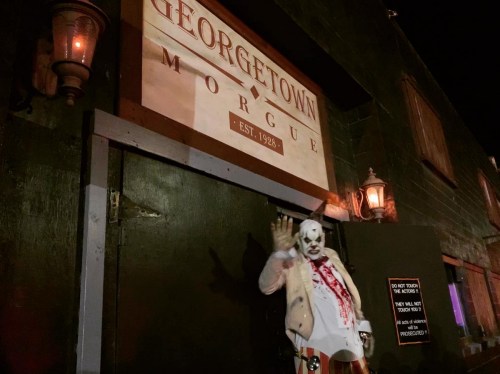 A creepy clown with fake blood stands outside the Georgetown Morgue, a dark building with a vintage sign.