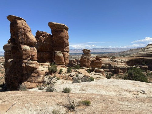 Rock formations rise against a clear blue sky, with distant mountains visible in the background. Desert landscape surrounds them.