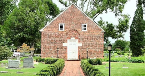 A brick building with a white door, surrounded by neatly trimmed hedges and gravestones in a serene garden setting.