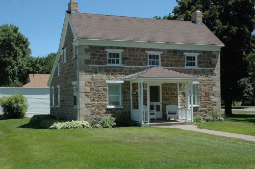A charming stone house with a front porch, surrounded by a well-maintained lawn and trees on a sunny day.