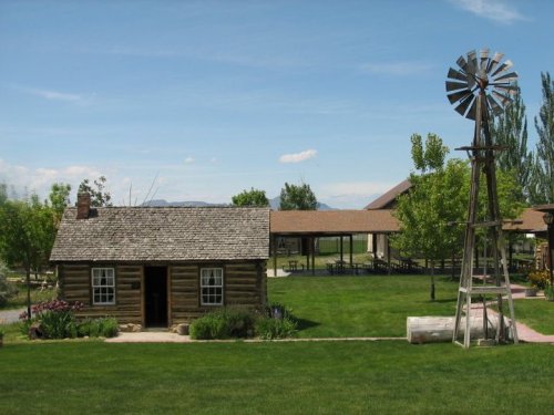 A rustic log cabin with a windmill nearby, surrounded by green grass and trees under a clear blue sky.