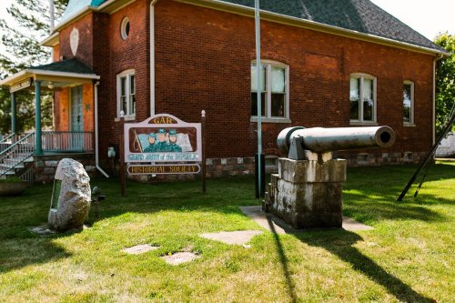 A historic building with a sign and a cannon displayed on the lawn, surrounded by grass and trees.