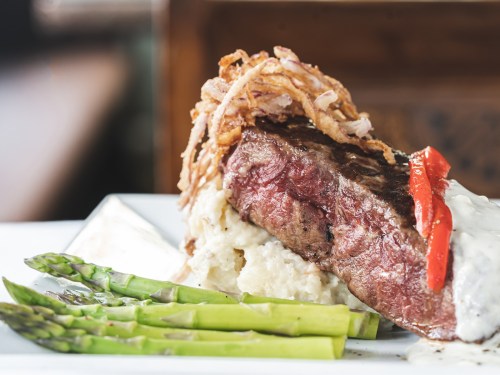 A plated dish featuring a juicy steak topped with crispy onions, served with mashed potatoes, asparagus, and red pepper.