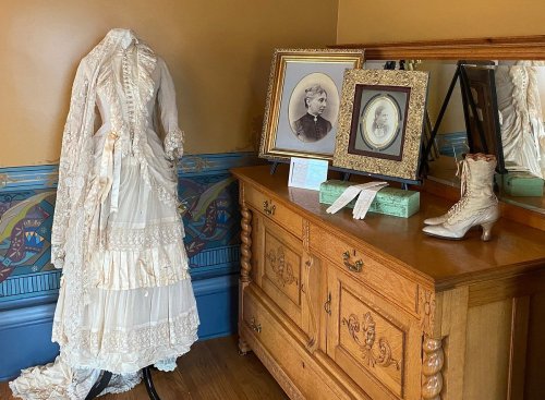 A vintage white dress on a mannequin beside framed portraits and an antique wooden dresser with a decorative boot.