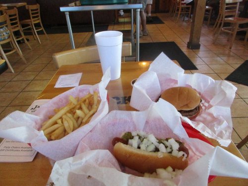 A table with a burger, hot dog topped with onions, fries, and a large drink in a casual dining setting.