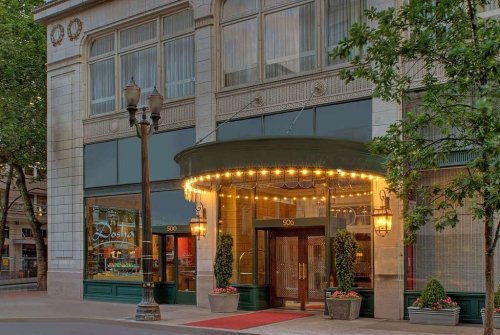 Elegant hotel entrance with marquee lights, potted plants, and a red carpet, framed by large windows and trees.