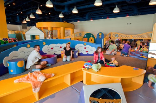 A colorful indoor play area with children and adults engaging in activities, surrounded by soft play structures and toys.