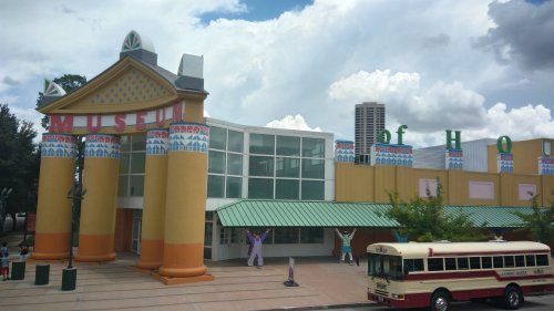 Colorful museum building with large columns, a green awning, and a vintage bus parked in front under a cloudy sky.