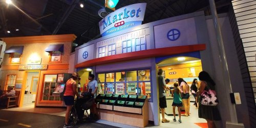 Children and adults explore a colorful market exhibit in a playful indoor setting.