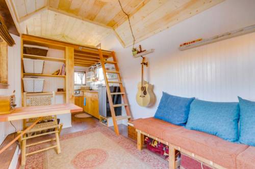 Cozy interior of a tiny house featuring a wooden ladder, kitchen area, and a seating area with blue cushions.