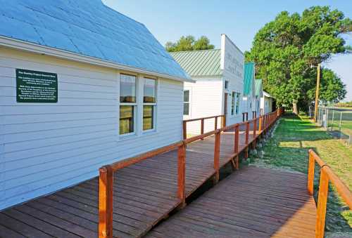 A wooden walkway leads to white buildings with metal roofs, surrounded by greenery and a clear blue sky.