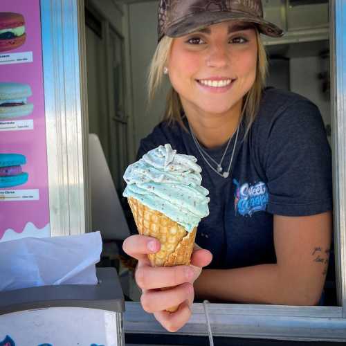 A smiling person holds a cone of blue mint ice cream in front of a food truck.