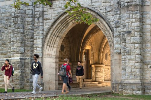 Students walk through a stone archway on a college campus, surrounded by greenery and historic architecture.