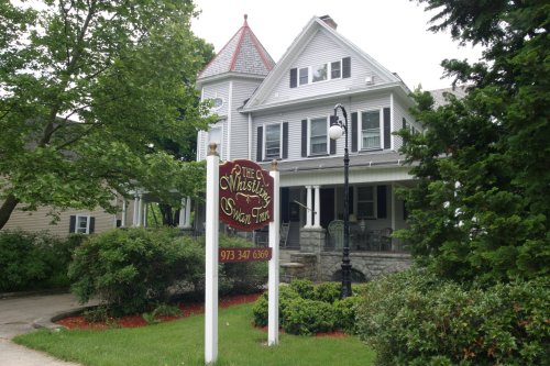 A charming gray house with a red roof and sign reading "The Whistling Swan Inn," surrounded by greenery.