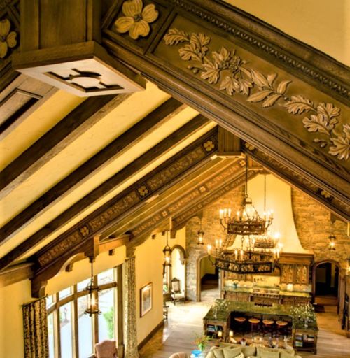 A spacious, elegantly designed room with wooden beams, ornate carvings, and a large chandelier above a kitchen area.