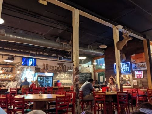 Interior of a rustic restaurant with wooden decor, a TV, and patrons dining at tables. A deer head is mounted on the wall.