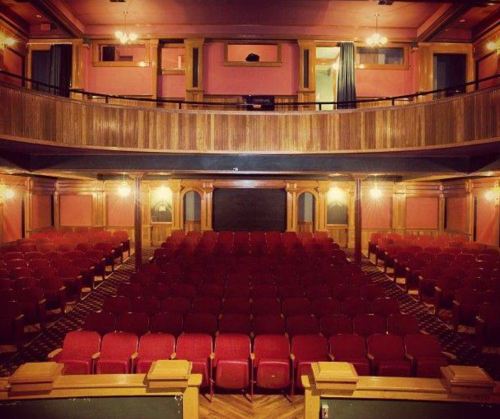 Interior of a vintage theater featuring red seats, wooden accents, and a balcony overlooking the stage area.