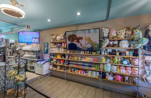 A cozy anime café featuring shelves of plush toys, snacks, and a large screen displaying anime characters.