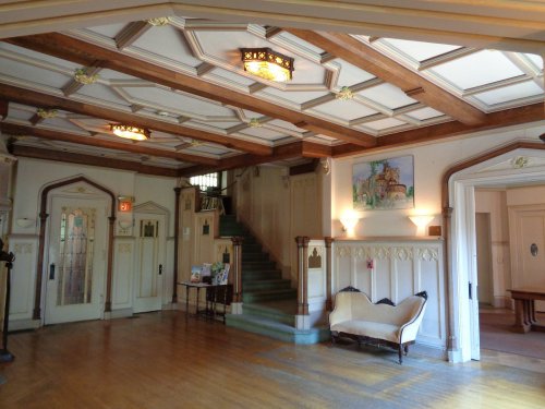 Elegant foyer with a wooden ceiling, staircase, and vintage furniture, featuring decorative lighting and artwork.