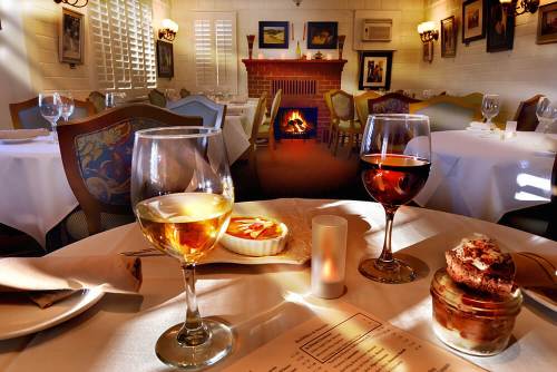 A cozy restaurant table set with white wine, red wine, and dessert, with a warm fireplace in the background.