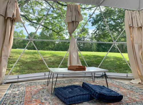 A cozy geodesic dome interior with a table, cushions, and large windows showcasing a green landscape outside.