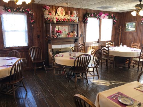 Cozy dining room with wooden walls, decorated with flowers, featuring round tables and a fireplace.