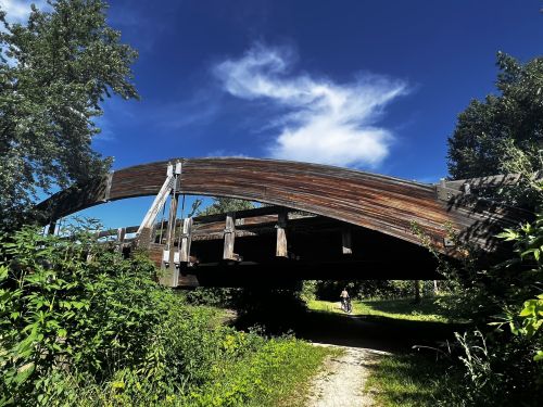 A wooden arch bridge surrounded by greenery under a blue sky with clouds, with a person biking in the distance.