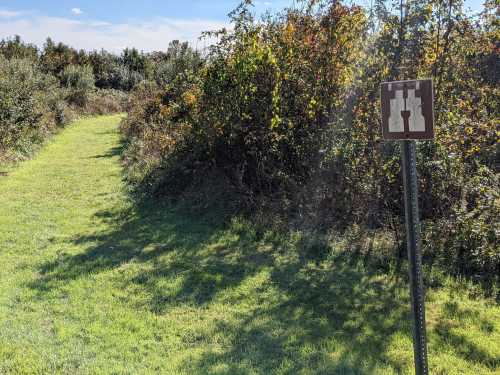 A grassy path leads through dense foliage, with a sign featuring two bottles on a post.