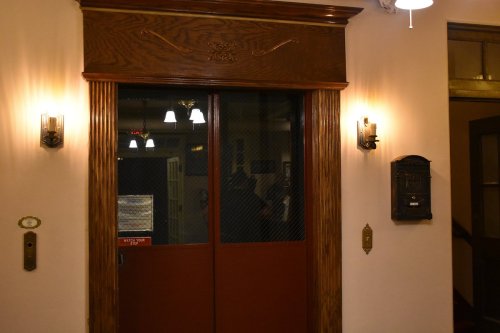 A vintage elevator door with wooden trim, flanked by wall sconces and a mail slot, in a softly lit hallway.