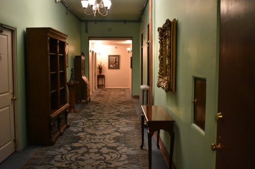 A long hallway with green walls, ornate decor, and a patterned carpet, featuring doors and decorative furniture.