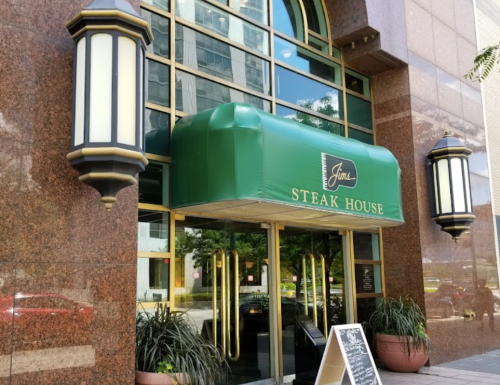 Entrance of a steakhouse with a green awning, large glass doors, and decorative light fixtures on a sunny day.