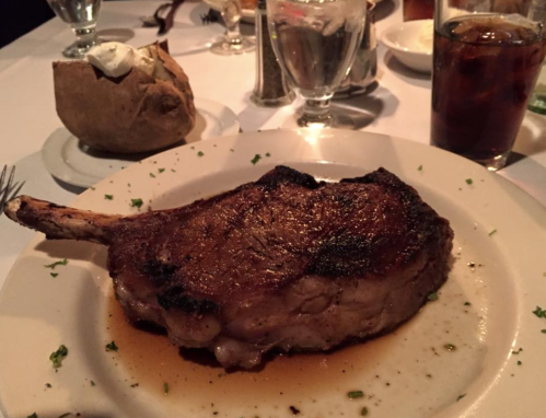 A juicy, grilled pork chop on a plate, garnished with herbs, alongside a baked potato and a glass of cola.