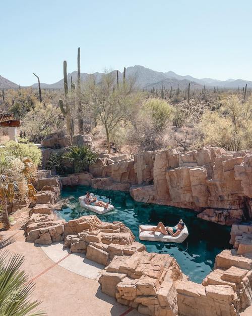 Two people relax on floating loungers in a rocky pool surrounded by desert scenery and cacti under a clear blue sky.