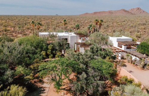 Aerial view of a modern white house surrounded by lush greenery and desert landscape with mountains in the background.