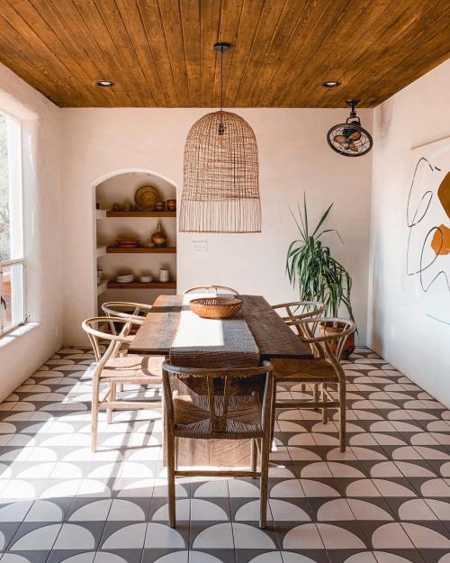 A bright dining area with a wooden table, woven pendant light, and patterned tile floor, featuring plants and decor.